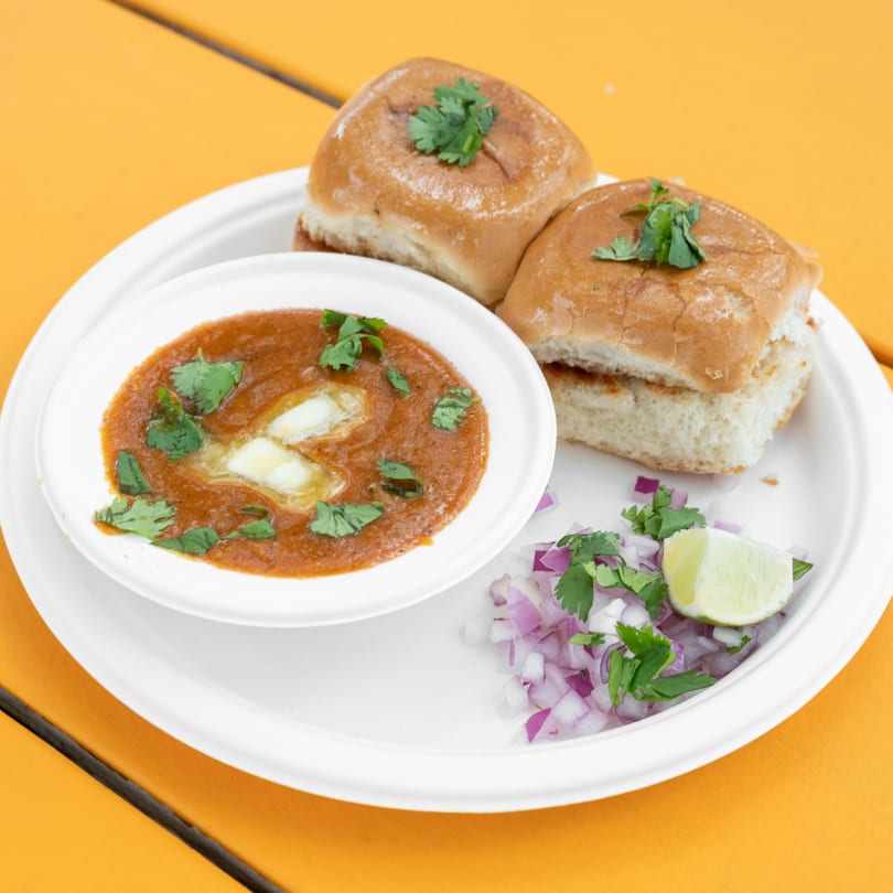 Pav Bhaji with two buttered buns, a bowl of vegetable curry, chopped onions, and a lime wedge.