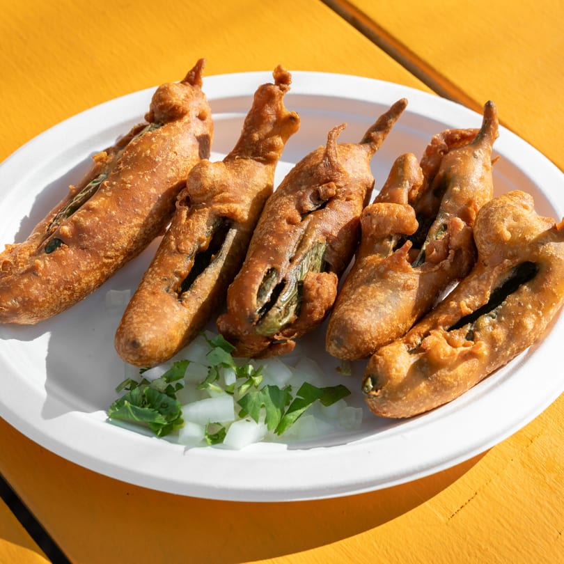 Six golden-brown fried chili peppers on a white plate with chopped onions and cilantro.