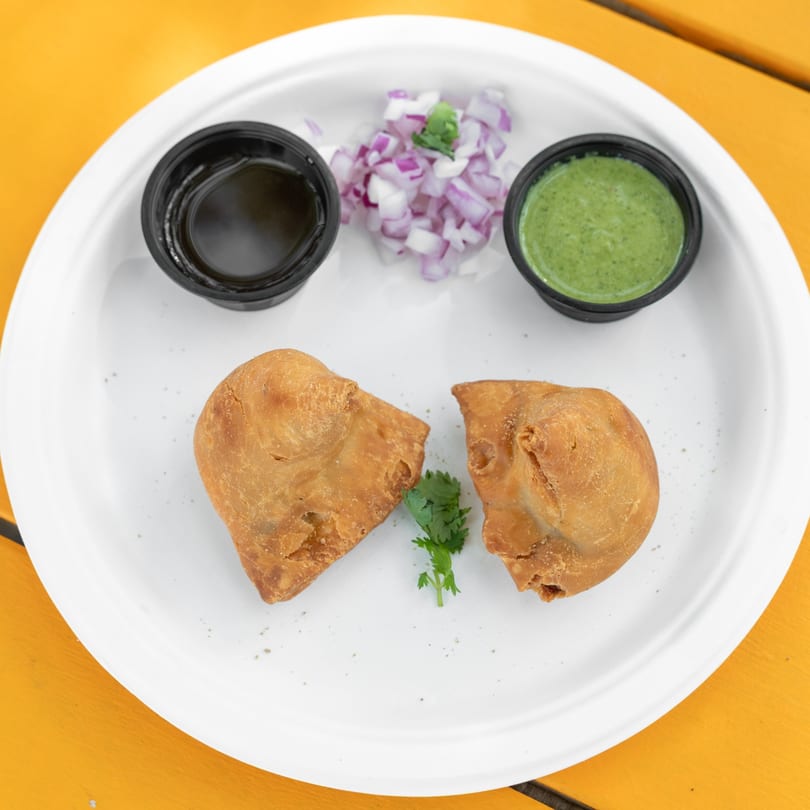 Two golden-brown samosas with cilantro, chopped red onions, and two dipping sauces on a white plate.