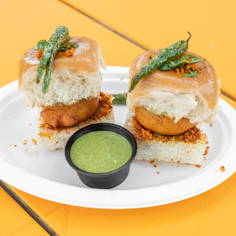 Two vada pavs with green chilies and a side of green chutney on a white plate.