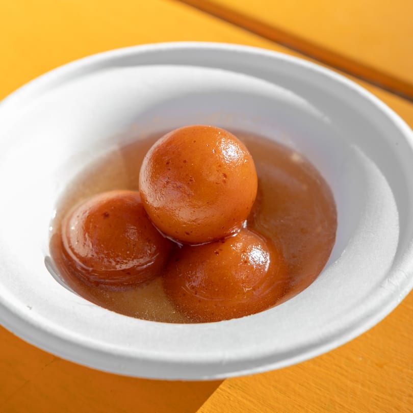 Three gulab jamun in a white bowl, soaked in a sweet, amber syrup on a yellow table.