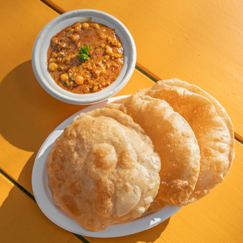 A bowl of chana masala with three fluffy puri breads on a bright yellow table.